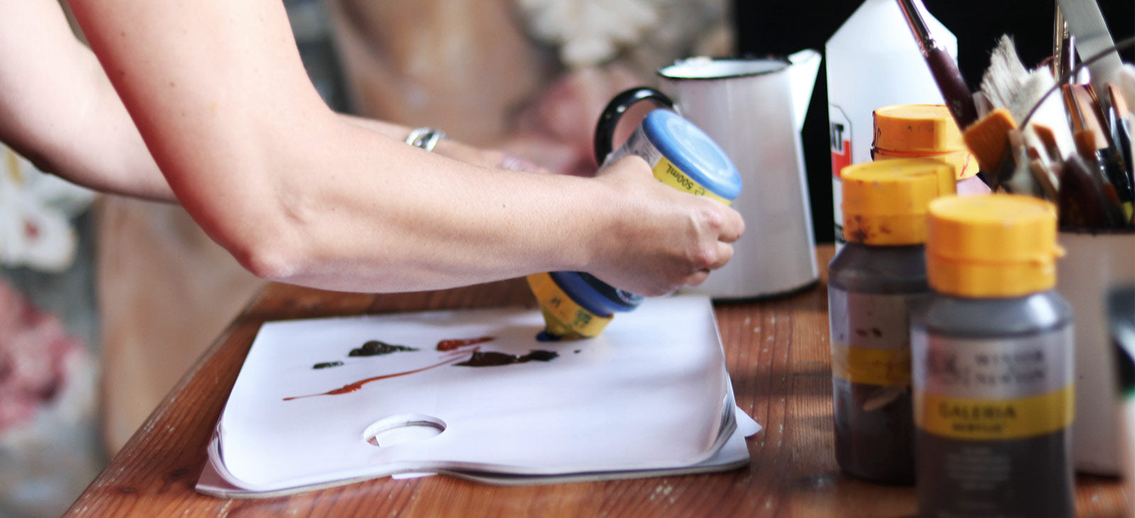 Artist’s hands mixing paint on a palette, with paint bottles and brushes on a wooden table.